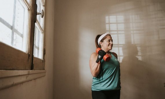 Senior woman in a brightly lit room exercises with dumbbells, promoting fitness at home.