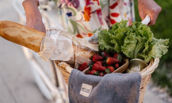 A woman in a floral dress holds a basket filled with bread, strawberries, and lettuce on her bicycle.